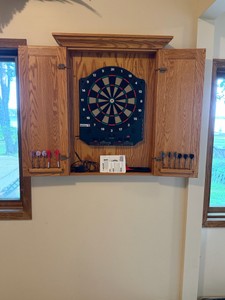 wall-mounted wooden dartboard cabinet, featuring classic raised-panel oak doors and a molded crown top. The cabinet houses a Sportcraft electronic dartboard inside, complete with pre-drilled dart storage built directly into the open left door panel.