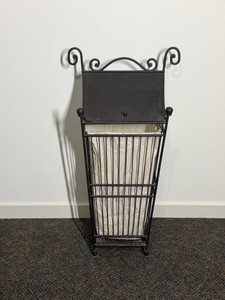 Vintage-style wrought iron laundry hamper equipped with a matching metal lid, decorative scrollwork handles, and a removable canvas liner bag.