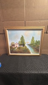 Framed landscape artwork appears to be a vintage or amateur oil painting on canvas, featuring a pastoral lakeside cottage scene with tall pine trees and soft, muted skies