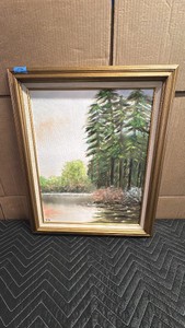 Framed landscape painting features a serene river or lake scene bordered by tall evergreen pine trees and soft pastel foliage, housed in an ornate gold-toned frame.