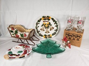 Christmas-themed items including Fitz and Floyd Santa plate, green glass tree dish, decorated sleigh basket, holiday mugs, bird figurine, and decorative plate with bells.