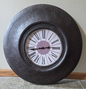 Large Wall Clock with Roman Numeral face with purple center. The clock is 24 round.&nbsp Frame features a textured, black, leather-like appearance.&nbsp Clock is battery operated.