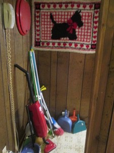Basement corner contents includes brooms, stick vac, a decorative toilet seat and a latch hook rug