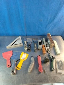 A variety of hand tools laid out on a metal surface against a blue background, mostly consisting of scraping, cutting, and marking tools.