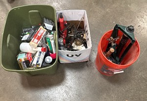Green tote of shims, nails, staples, spray paint, plastic wrap. Orange bucket  with 2 sprinklers and hinge. White box with garage items